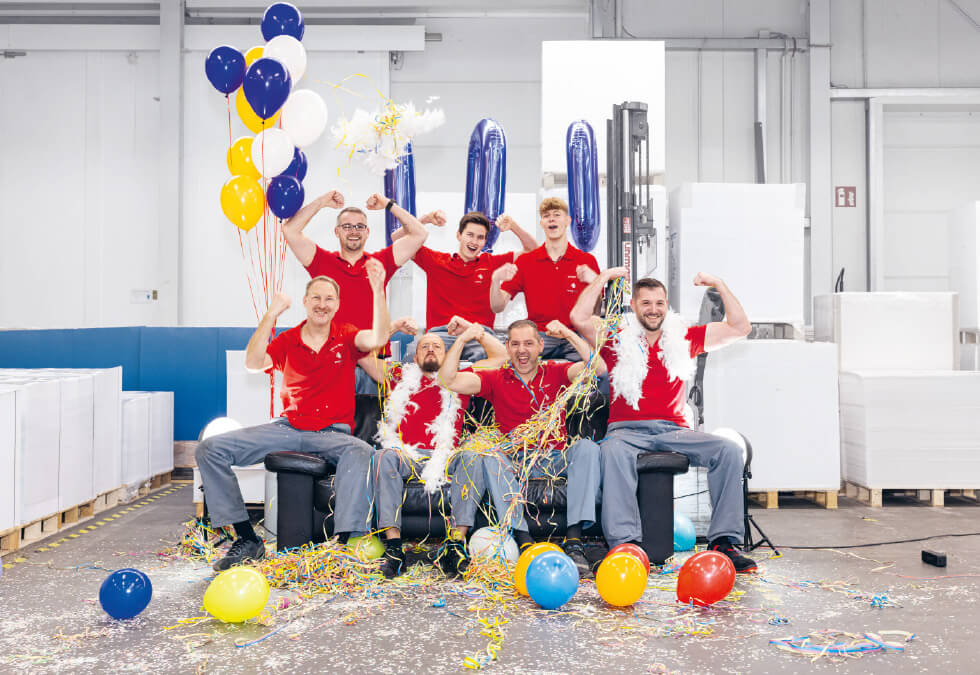 Beisner_1 - Beisner Druck GmbH & Co. KG Sieben Menschen in roten Hemden und grauen Hosen feiern fröhlich in einer Lagerhalle, sitzen auf schwarzen Stühlen zwischen Luftballons, Luftschlangen und Konfetti - ein weiterer Festtag in der Druckerei Hannover. Einige fuchteln mit den Armen, um die Stimmung noch zu steigern.
