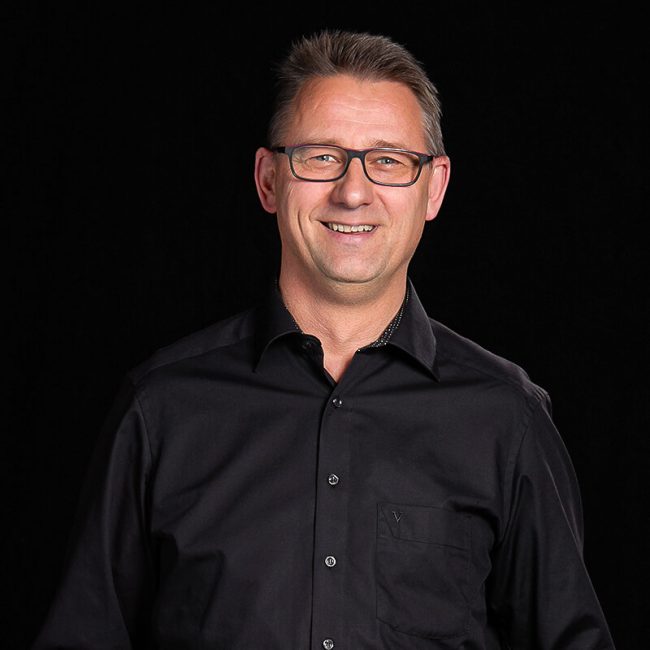 Team_Eckhardt-Schulz_web-650x650 - Beisner Druck GmbH & Co. KG Ein Mann mit kurzen braunen Haaren und Brille lächelt, während er vor einem schwarzen Hintergrund steht. Er trägt ein schwarzes Button-up-Hemd, dessen oberster Knopf geöffnet ist und das das sympathische Gesicht der Druckerei Bremen darstellt.