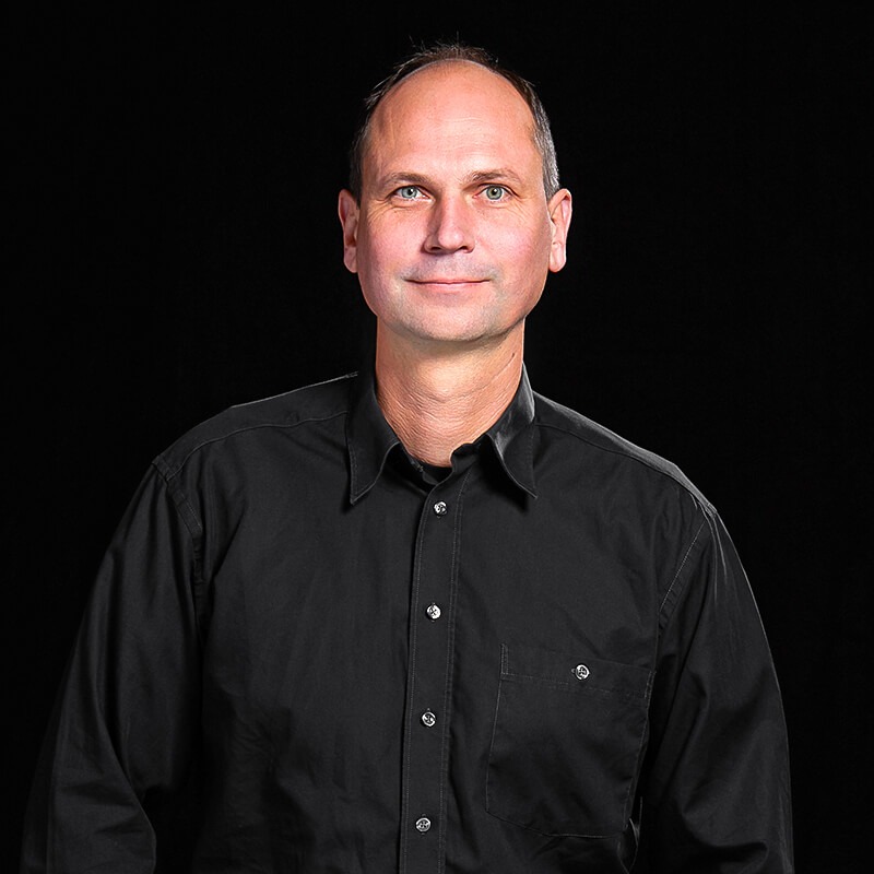Team_Helge_Beisner_web - Beisner Druck GmbH & Co. KG Ein Mann mittleren Alters mit kurzen Haaren und schwarzem Button-up-Hemd steht vor einem schlichten schwarzen Hintergrund und schaut mit einem leichten Lächeln direkt in die Kamera - professionell und sympathisch, perfekt für eine Teamvorstellung der Druckerei Hannover.