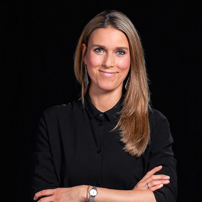 Team_Julia_Roggendorf_web-650x650 - Beisner Druck GmbH & Co. KG Eine Frau mit langen, glatten blonden Haaren und einem schwarzen Button-up-Hemd steht lächelnd mit verschränkten Armen vor einem schwarzen Hintergrund. Sie trägt eine Uhr und Ohrringe - perfekt für ein Teamfoto in der Druckerei Hamburg oder bei Offsetdruck Hamburg.