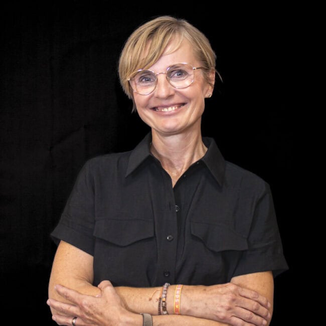 Team_Nela_Janssen_web2-650x650 - Beisner Druck GmbH & Co. KG Eine lächelnde Frau mit kurzen blonden Haaren und Brille steht vor einem schwarzen Hintergrund, trägt ein kurzärmeliges schwarzes Hemd und hat die Arme verschränkt - perfekt für ein Teamporträt der Druckerei Hamburg.