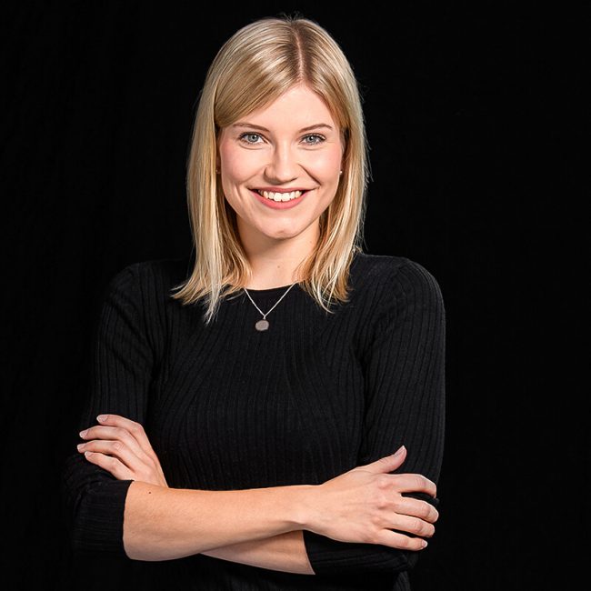 Team_Sissy_Steffens_web-650x650 - Beisner Druck GmbH & Co. KG Eine lächelnde Frau mit glattem blondem Haar, die ein schwarzes langärmeliges Oberteil und eine Halskette mit Anhänger trägt, steht mit verschränkten Armen vor einem schlichten schwarzen Hintergrund und verkörpert den professionellen Geist der Druckerei Hamburg.