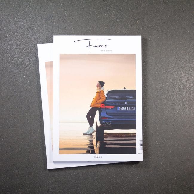Eine Zeitschrift mit dem Titelbild einer Person in orangefarbener Jacke und Mütze, die sich vor einem Sonnenuntergang an ein blaues Auto lehnt, wird auf eine andere ähnliche Zeitschrift auf einer grauen Fläche gelegt.