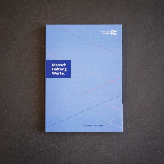 Ein blaues Büchlein mit geometrischen Linien auf einer dunklen Oberfläche. Auf dem Umschlag steht "Mensch. Haltung. Werte." in einem blauen Kasten und "Geschäftsbericht 2018" am unteren Rand; das Logo der "Deutsche Wohnen" befindet sich in der oberen rechten Ecke.