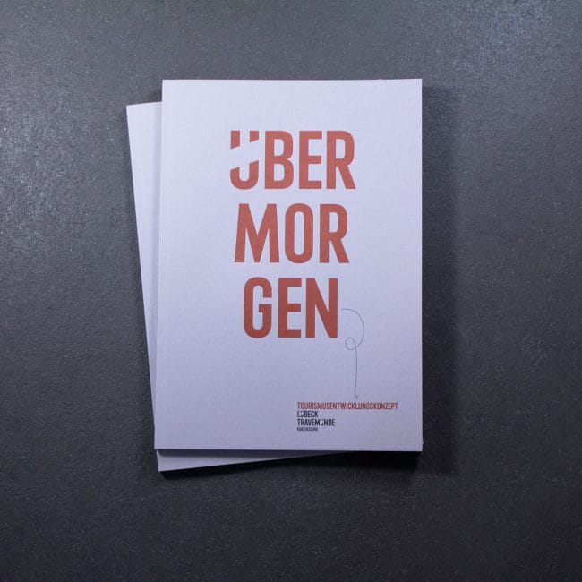 Ein weißes Heft mit dem Wort ÜBERMORGEN in fetten orangefarbenen Buchstaben liegt auf einer grauen Fläche, darunter ist ein weiteres ähnliches Heft teilweise sichtbar.