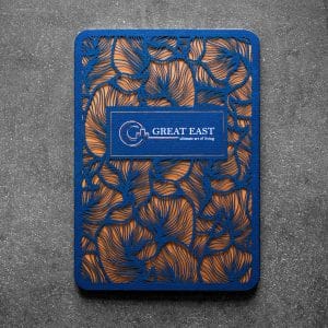 Eine rechteckige Karte mit blauem und goldenem Blattmuster und dem Text GREAT EAST - classic art of living" auf einem blauen Etikett in der Mitte, vor einem dunklen, strukturierten Hintergrund.
